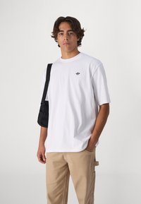 White cotton t-shirt with short sleeves, featuring a small black logo on the chest. Paired with beige cargo pants and a black bag.