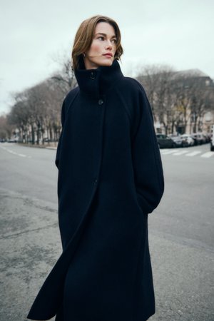 Donna con capelli castani corti che indossa un lungo cappotto scuro con colletto alto, si trova su una strada cittadina con alberi spogli e auto parcheggiate.