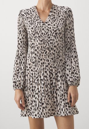 Woman wearing a long-sleeve, pleated, V-neck beige dress with black abstract spots and elastic cuffs, standing against a white background.