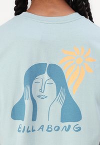 Lys blå bomulls T-skjorte med et frontgrafikk av en kvinne med langt hår og en sol, trykket i mørkeblå og oransje farger.