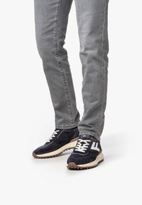 Gambe che indossano jeans grigi e sneakers in pelle scamosciata nera con lacci bianchi e un logo "F" bianco, in piedi su uno sfondo bianco.