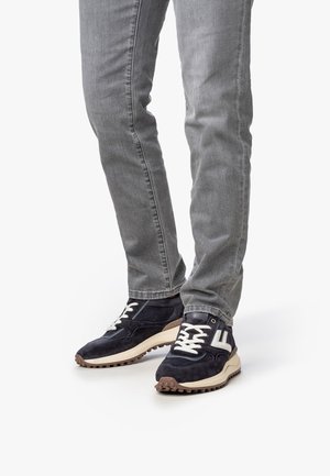 Gambe che indossano jeans grigi e sneakers in pelle scamosciata nera con lacci bianchi e un logo "F" bianco, in piedi su uno sfondo bianco.