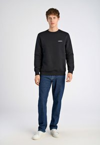 Svart sweatshirt med rund hals och ribbad mudd, ihop med blå denimjeans och vita sneakers. Sweatshirten har en liten logotyp.