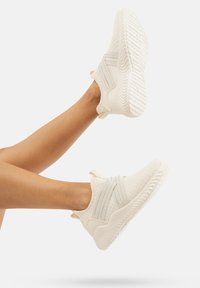 Zapatos deportivos blancos con un upper de tela texturizada, que cuentan con dos bandas elásticas grises en la parte delantera y una suela de goma estriada.