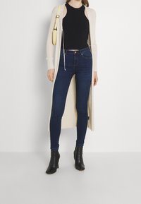 Beige long cardigan over a black sleeveless top, paired with dark blue skinny jeans and black heeled ankle boots. Smooth knit texture visible.