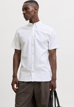 Hombre con una camisa blanca de manga corta abotonada y pantalones marrones, sosteniendo un bolso negro, mirando hacia un lado.