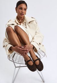 Ruskeat leopardi-kuosiset Mary Jane -kengät, joissa on remmi, pehmeää kangasta, pyöreä kärki. Malli istuu jalat ristissä langan tuolilla, päällään beige trench coat.