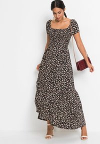 Robe maxi noire avec un petit motif floral rose et blanc, décolleté carré, corsage ajusté, jupe évasée. Modèle tenant un sac bordeaux.