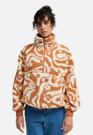 Kobieta ubrana w polarowy pulower w odcieniach beżu i bieli z półzamkiem i wysokim kołnierzem, w połączeniu z niebieskimi dżinsami, na jednolitym tle.