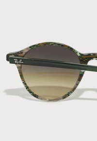 Ochelari de soare rotunzi cu ramă din chihlimbar verzuie, lentile cu un gradient de la maro la bej deschis și un braț verde cu logo-ul Ray-Ban.