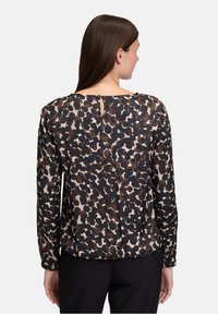 Frau mit langen braunen Haaren, die eine Bluse mit langen Ärmeln im schwarz-braunen Leopardenmuster trägt, von hinten vor einem schlichten Hintergrund betrachtet.