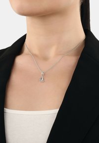 Sölvhalssmycke med en segelbåtsberlock med detaljer och en fin kedja. Bärs över en ljus topp, mot en svart jacka.