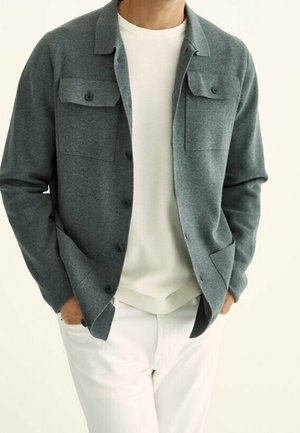 Homme portant une veste grise boutonnée avec des poches à rabat sur une chemise blanche et un pantalon blanc, les mains dans les poches de la veste.