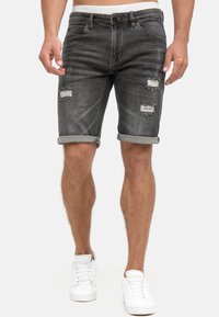 Pantalones cortos de mezclilla oscuros con desgastes y dobladillos deshilachados. Puños enrollados y cierre de cremallera. Combinados con zapatillas blancas. Diseño casual.