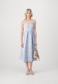 Robe florale bleu clair avec des fines bretelles, silhouette A-line fluide et taille froncée. Assortie à un sac à main beige et des sandales.