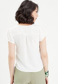 Haut blanc à manches courtes avec une coupe décontractée, présentant un détail de pli au dos, une texture lisse et un tissu léger, associé à un pantalon vert olive.