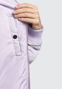 Hele lilla bomber-jakk, millel on sile ja läikiv tekstuur. Omab soonikpihte, lukuga sulgemist ja nööbiga taskute detaili.