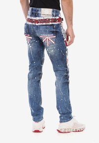 Jeans en denim bleu foncé avec des motifs de éclaboussures blanches. Présente des patchs Union Jack sur les poches et une ceinture texturée avec des lettres rouges.