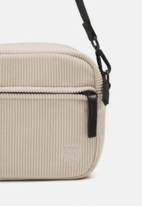 Borsa a tracolla in corduroy beige con superficie a coste testurizzata, dotata di una tasca con zip nera e tracolla regolabile. Emblema impresso sulla parte anteriore.