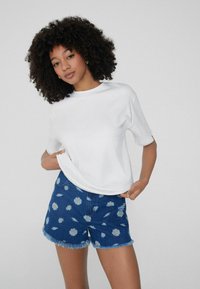 Weißes T-Shirt mit kurzen Ärmeln, kombiniert mit blauen Jeansshorts mit Margeritenmuster und fransigen Säumen, die einen lässigen Sommerlook kreieren.