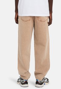 Μπεζ τζιν παντελόνια με φαρδύ πόδι και υφασμάτινη υφή, με πίσω τσέπες και γυρισμένα τελειώματα, σε συνδυασμό με μαύρα και ασημένια αθλητικά παπούτσια.