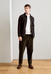 Camisa negra de botones sobre una camiseta blanca, combinada con pantalones de pana verde oscuro y botines marrones, de pie sobre un suelo de madera.
