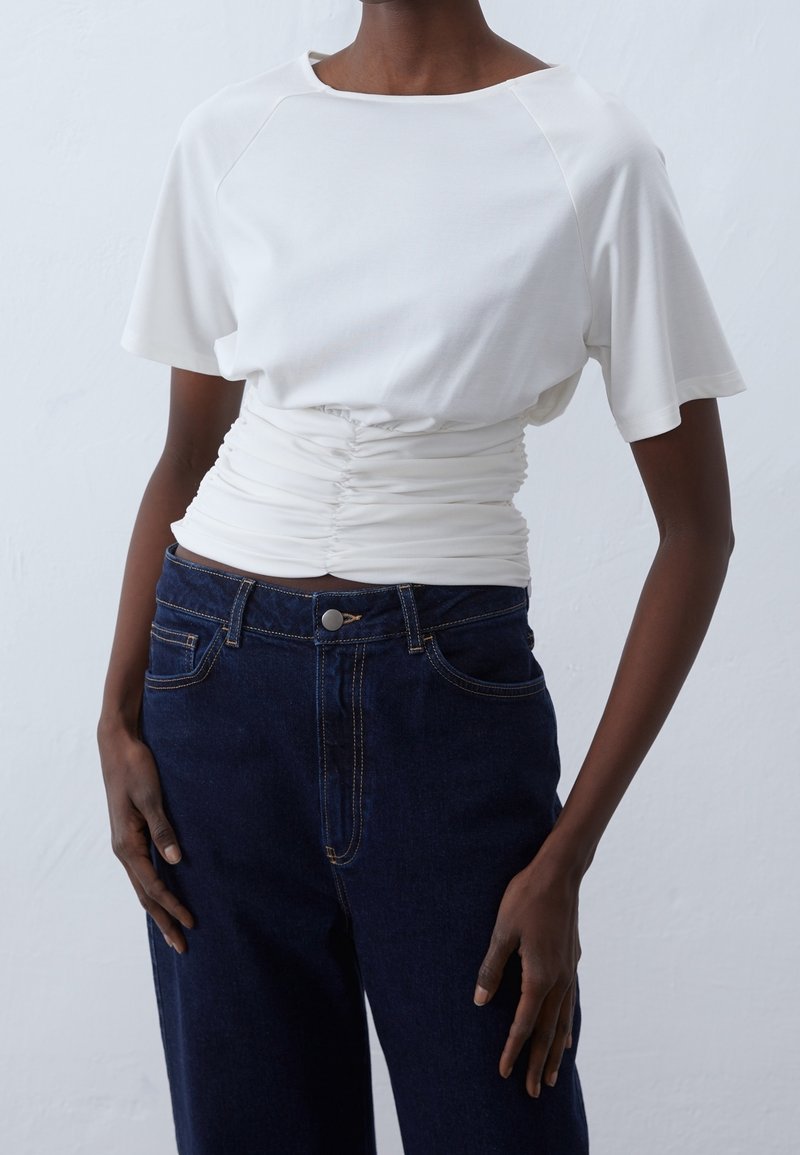 Personne portant un haut blanc à manches courtes froncé rentré dans un jean bleu foncé taille haute, debout devant un fond clair uni.