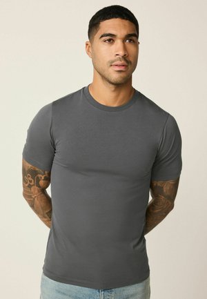 Rövid hajú, karját tetoválások díszítik, szűk szabású szürke környakú pólót és világoskék farmert viselő férfi, egyszínű háttér előtt áll.