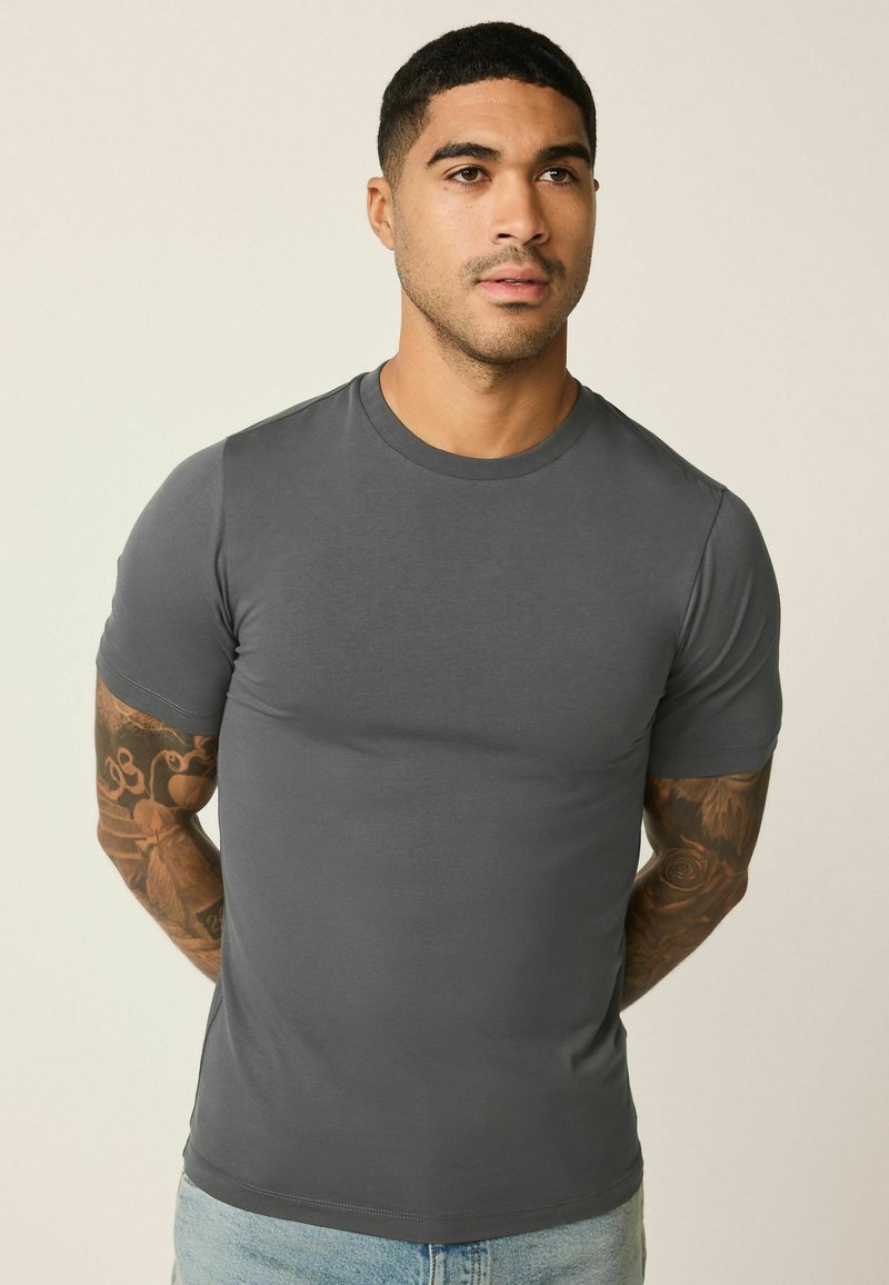 Mann mit kurzen Haaren und Armtattoos, der ein eng anliegendes graues T-Shirt mit Rundhalsausschnitt und hellblaue Jeans trägt, steht vor einem einfarbigen Hintergrund.