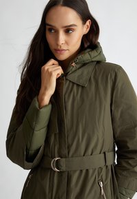 Olijfgroene pufferjack met een gestrikt middel, verstelbare capuchon en ritszakken aan de zijkant. Het heeft een gedempte glans en een gewatteerde textuur.