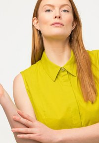 Blusa sin mangas con cuello en un vibrante verde lima. Presenta un botón decorativo en el escote. Tejido suave con una textura crujiente.