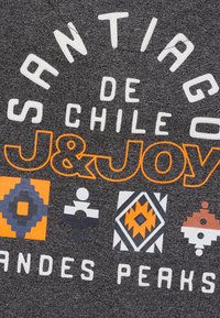 Donkergrijze stof met opvallende witte en oranje tekst die "Santiago de Chile" en "J&Joy" zegt, met geometrische patronen in verschillende kleuren eronder.