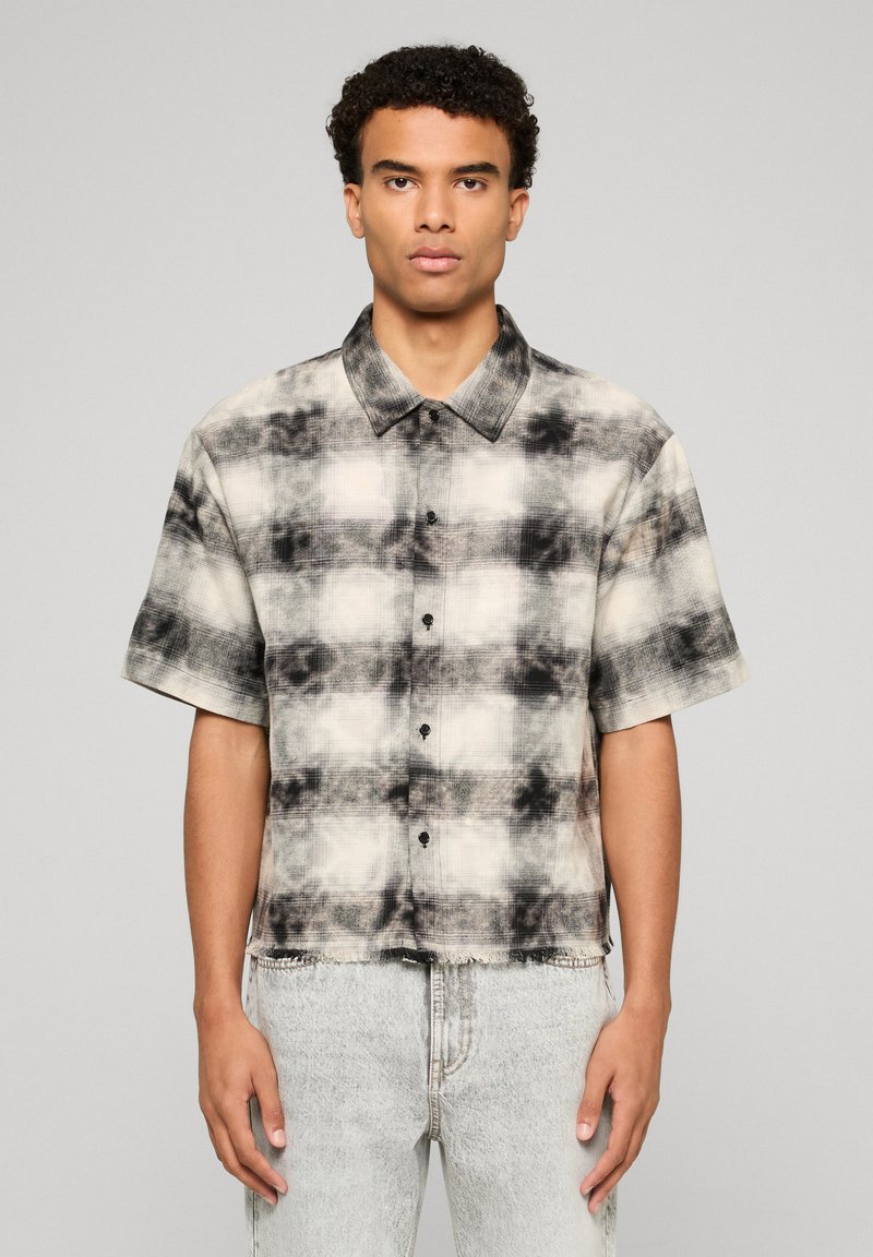 Jeune homme portant une chemise à carreaux noire et blanche à manches courtes et un jean clair, debout devant un fond gris uni.