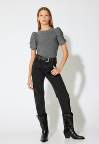 Grauer, kurzärmliger, strukturierter Oberteil; hochtaillierte schwarze Jeans; schwarze Cowboystiefel mit dekorativer Stickerei; stehend vor einem weißen Hintergrund.