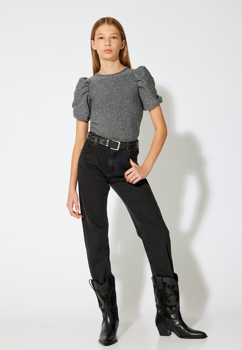 Grauer, kurzärmliger, strukturierter Oberteil; hochtaillierte schwarze Jeans; schwarze Cowboystiefel mit dekorativer Stickerei; stehend vor einem weißen Hintergrund.