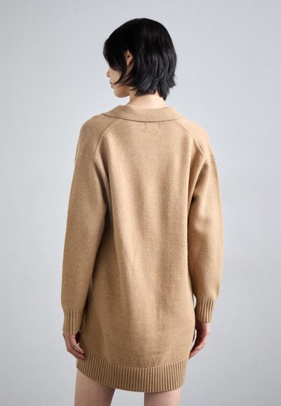 Beige gebreide jurk met een relaxte pasvorm, ribbels aan de manchetten en onderrand, en een zachte textuur. Heeft een brede kraag en een ontwerp met afhangende schouders.