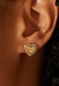Gouden hartvormige oorbellen met een "GUESS" gravure, omgeven door rhinestones. Ontworpen om in de oorlel te dragen, met een gladde textuur.