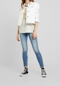 Witte denim jas, grijs graphic T-shirt, lichtblauwe skinny jeans en zwarte high-top sneakers. Eenvoudig ontwerp met een opvallende casual stijl.