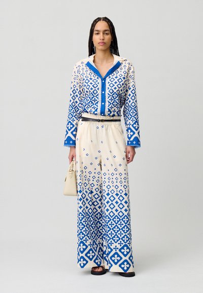 Camisa de manga larga azul y blanca con un patrón, detalles de botones y cuello, combinada con pantalones blancos holgados que presentan diseños geométricos azules.