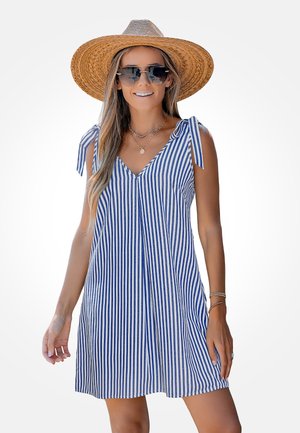 Frau trägt ein blau-weiß gestreiftes ärmelloses Kleid mit Schulterbändern, großen Strohhut, Sonnenbrille und mehrfachen Halsketten.