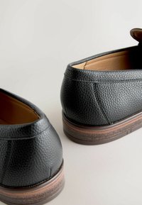 Mustad musta nahast kingad, millel on tekstureeritud pind, beež sisevooder ja madal pruun puidust konts, millel on peen aktsentliin.