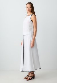 Haut sans manches en lin blanc avec une bordure noire, associé à une jupe maxi en lin blanc présentant une ligne verticale noire. Porté avec des sandales noires à lanières.