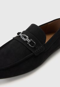 Mocasinés de ante negros con una textura suave y una tira decorativa que presenta herrajes plateados y un acento trenzado.