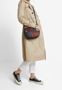 Kulatá taška s kombinací vínových, námořnických a černých kožených segmentů, s užším zipem a popruhem přes rameno, doplněná béžovým trenčkotem.