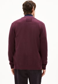 Pull-over en marron avec des manches longues, col et poignets côtelés, texture lisse, et un patch carré dans le haut du dos. Conception à ourlet plat.