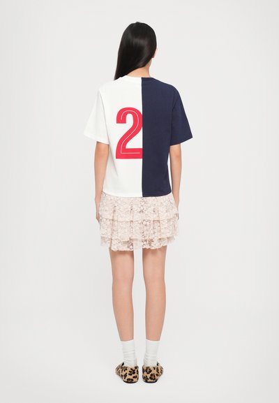 Een half witte en half marineblauwe t-shirt met een grote rode '2' op de achterkant, gecombineerd met een lichtroze geklede tiered rok en slippers met luipaardprint.