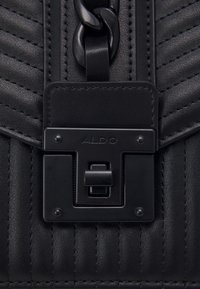 Svart vadderad konstläderväska med ett framträdande metallspänne, märkt med "ALDO." Har en dekorativ kedja och texturerad yta.