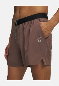 Rudos sportiniai šortai su juodu juosmeniu, raišteliu, šoninėmis kišenėmis ir perforuotais akcentais vėdinimui. Matomas Under Armour logotipas.