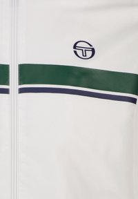 Weiße Jacke mit grünen und marineblauen Streifen über der Brust. Enthält ein kreisförmiges Logo. Strukturiertes Material mit einem Reißverschluss.