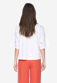 Bílé tričko s krátkými rukávy a zkráceným střihem, zdobené barevným výšivkou ve tvaru srdce v růžové, červené a zelené barvě.